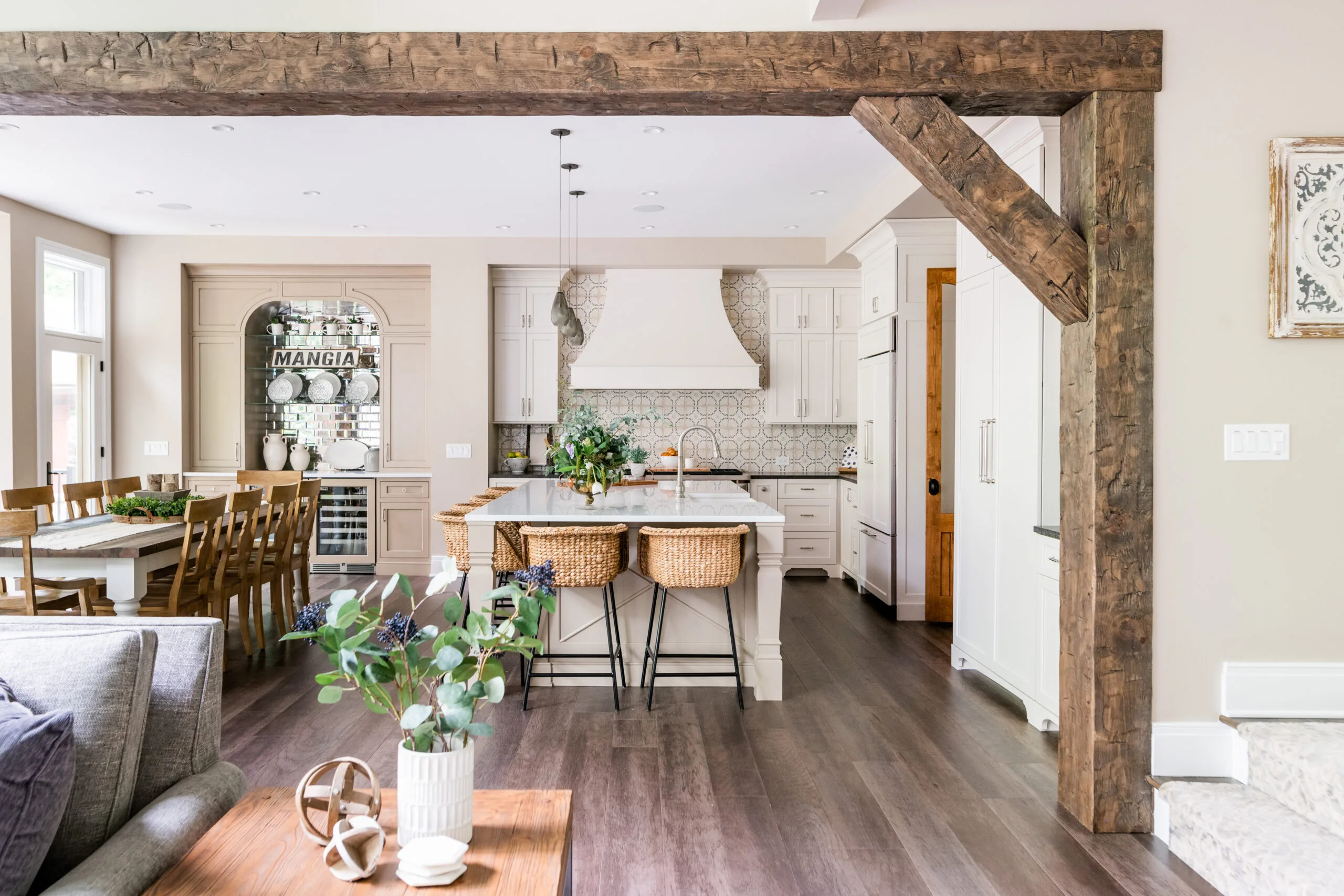 Beam wrapped opening between great room and kitchen. Custom luxury cabinetry and wood hood with painted cabinetry and rattan barstools