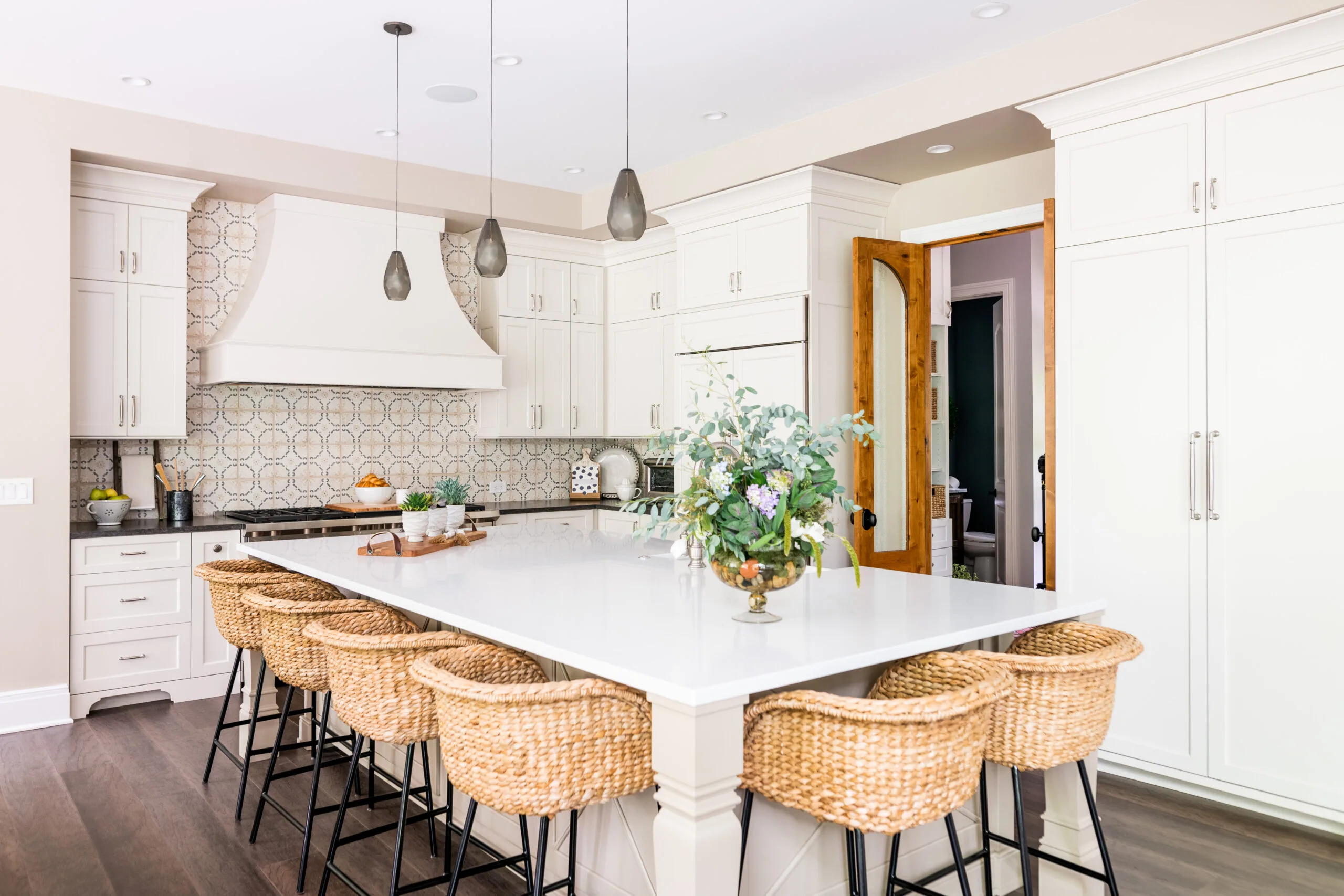 Beam wrapped opening between great room and kitchen. Custom luxury cabinetry and wood hood with painted cabinetry and rattan barstools