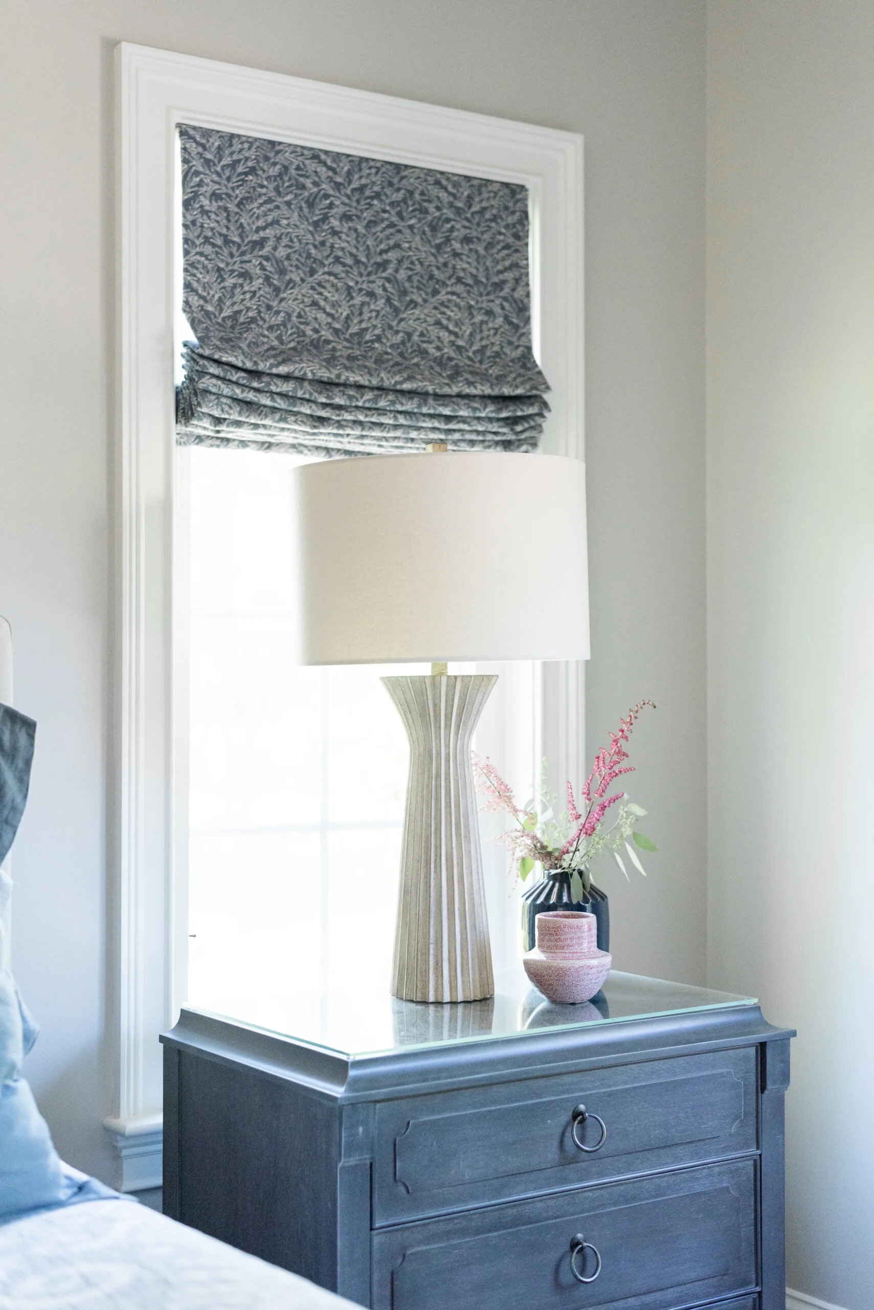Pale blue nightstand with a cream lamp in front of a window with a custom blue, patterned roman shade