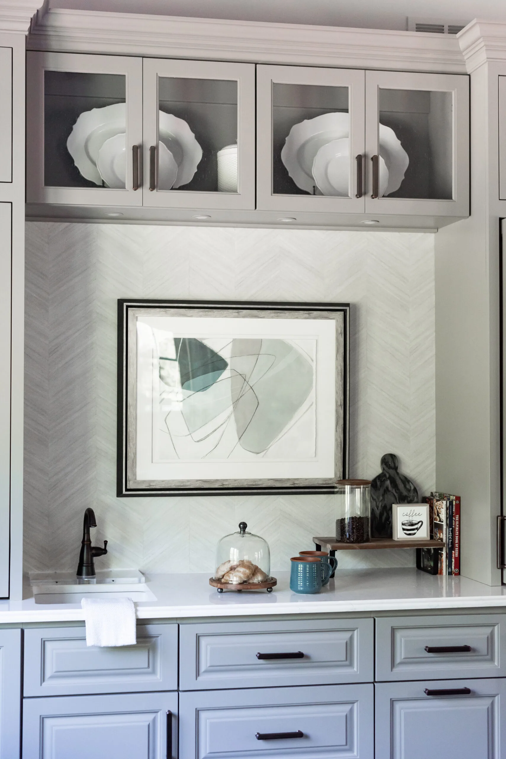 Wet bar with blue and neutral cabinetry, blue hued artwork above the countertop, coffee mugs, and small sink