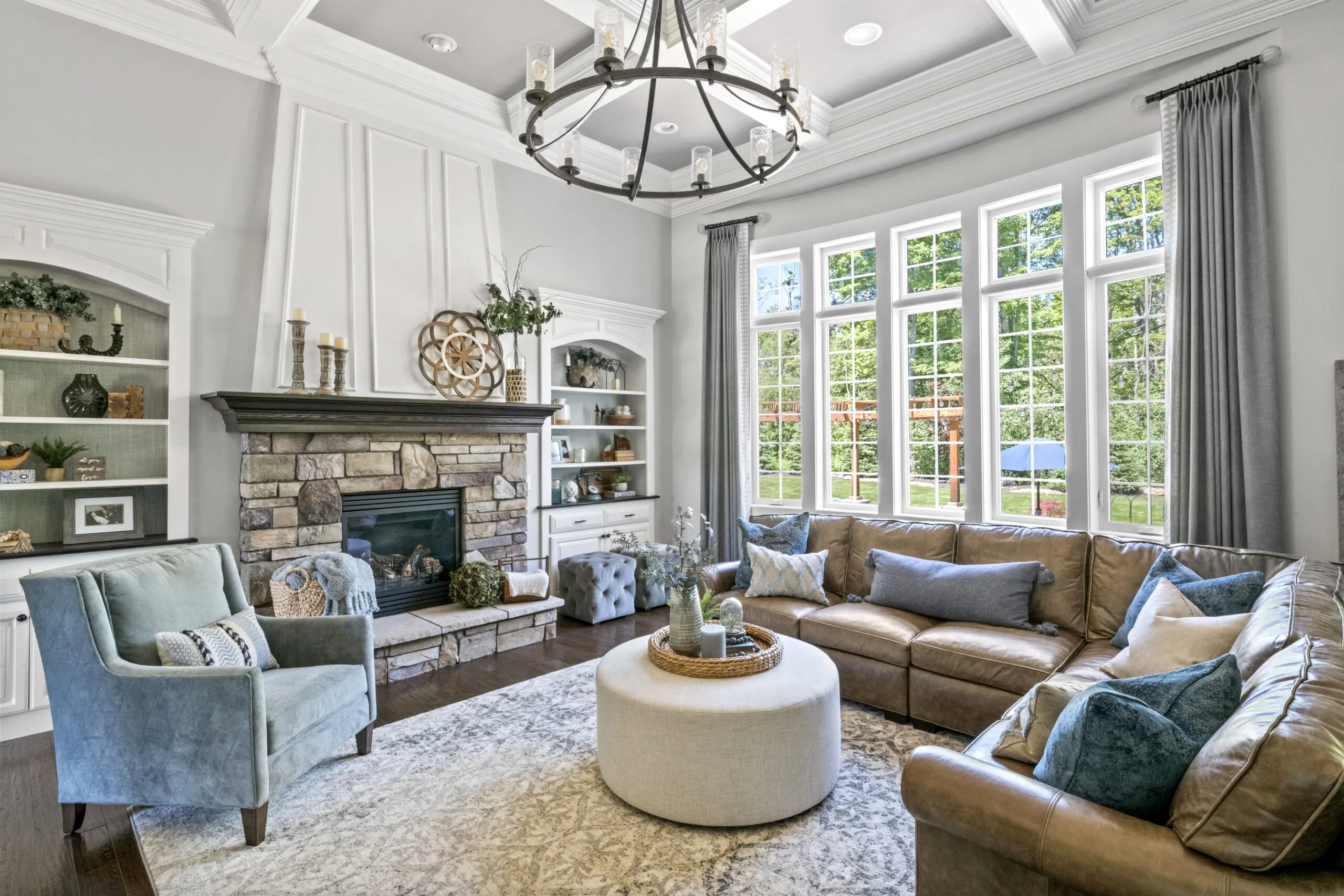 Overview of a living room with a large bank of windows, a stone fireplace, brown couch, round ottoman, and blue accent pieces