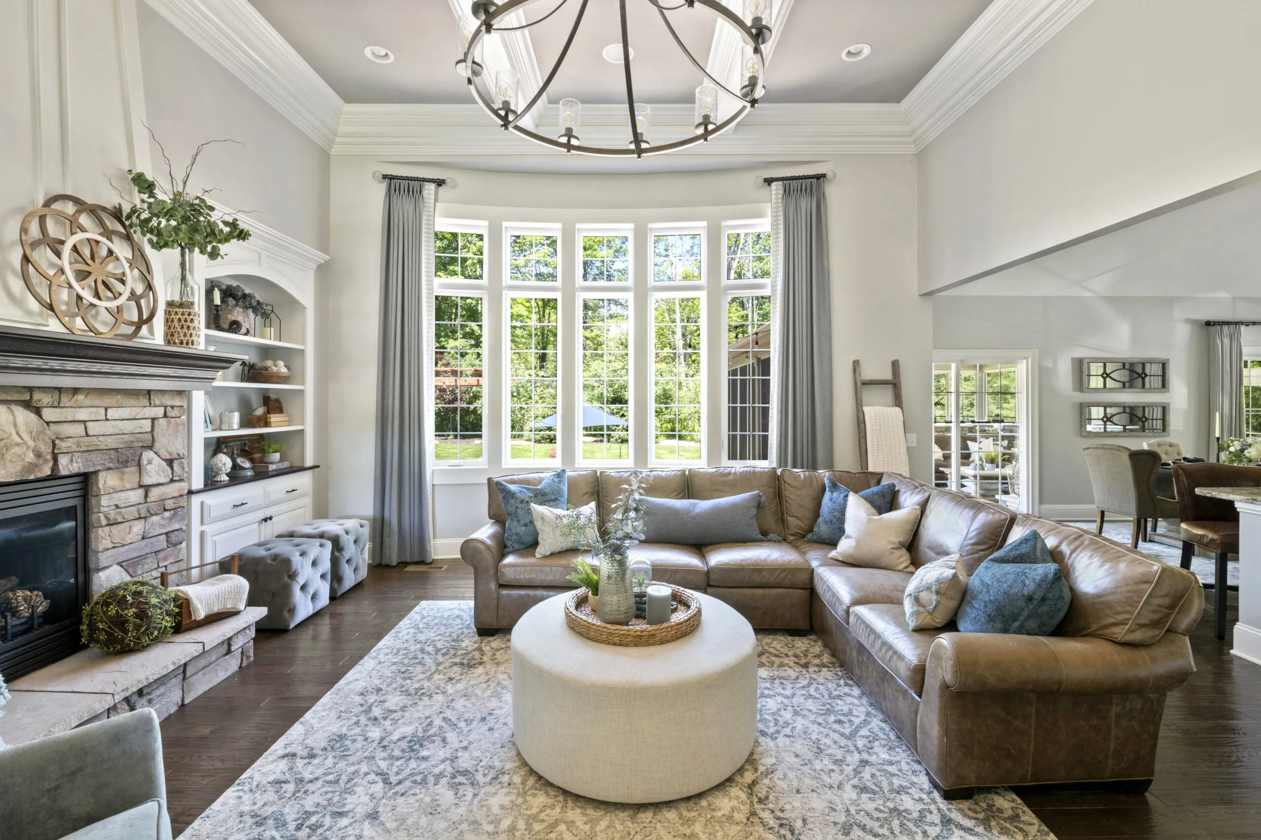 An overview of a living room with a large bank of windows, built-in cabinetry, brown couch, round ottoman, and blue accent pieces