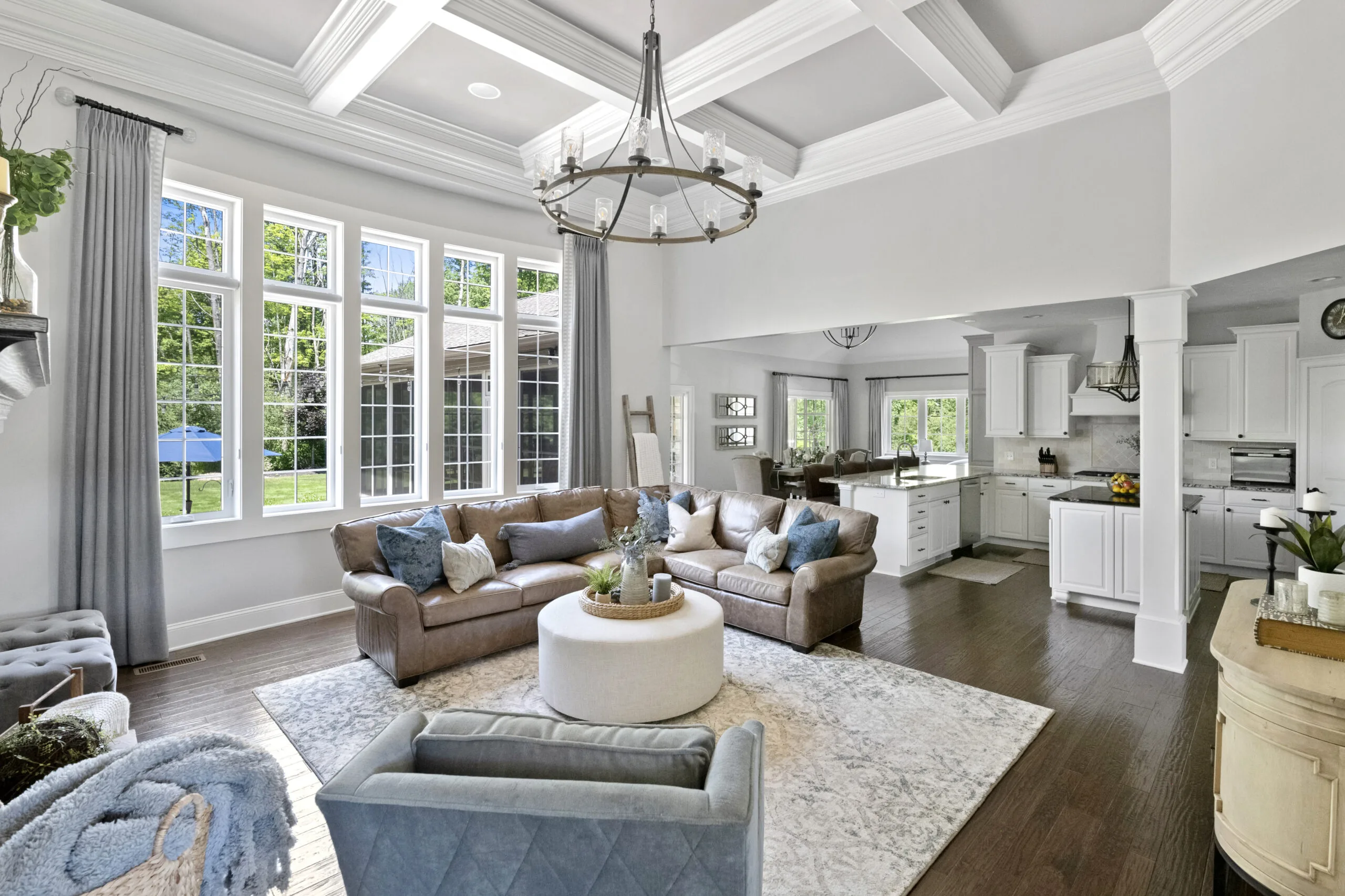 View of a living room with brown couch, round ottoman, and large bank of windows looking into a kitchen