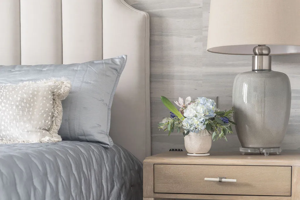 Close up image of a light wood nightstand next to an upholstered bed with florals and lamp sitting on top