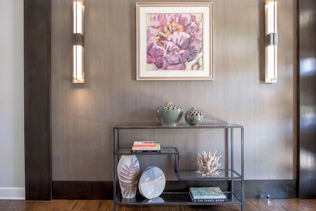 Metal and glass console table filled with accessories with purple artwork hanging above and long sconces on either side