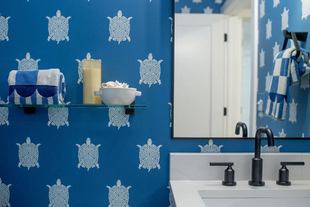 A floating glass shelf to the left of a bathroom sink and mirror with blue/white turtle wallpaper covering the walls