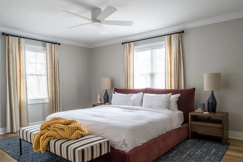 Bedroom with a deep red upholstered bed, a striped bench at the end of the bed, and side tables with blue lamps on top