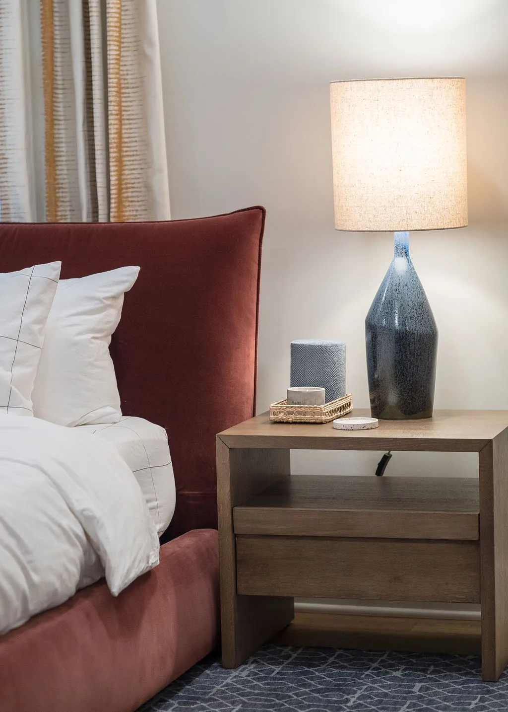 Image of the corner of a red, velvet headboard and a wooden side table and blue lamp to the right