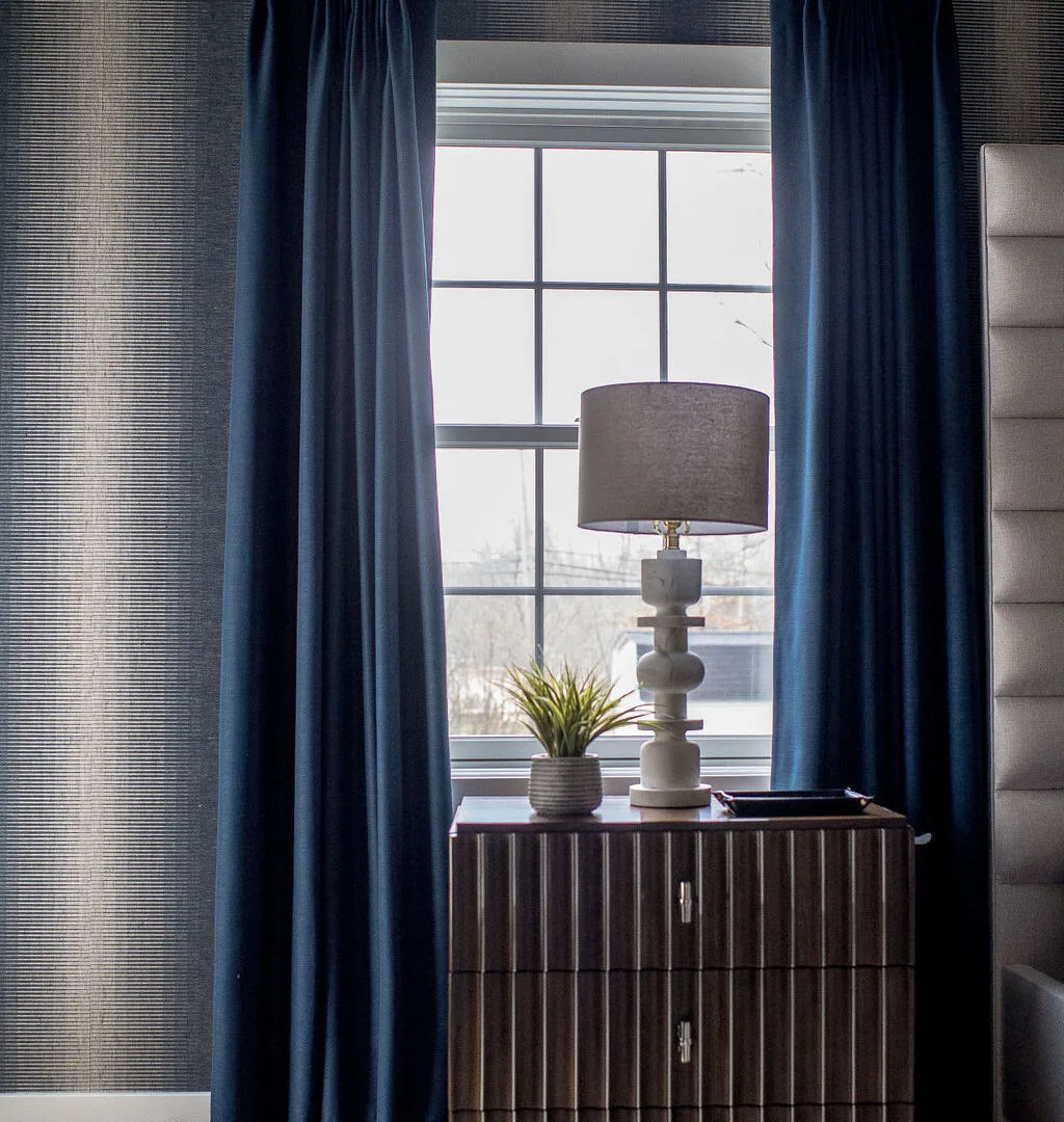 Table lamp sitting on top of a dark wooden nightstand in front of a window with blue panel drapes