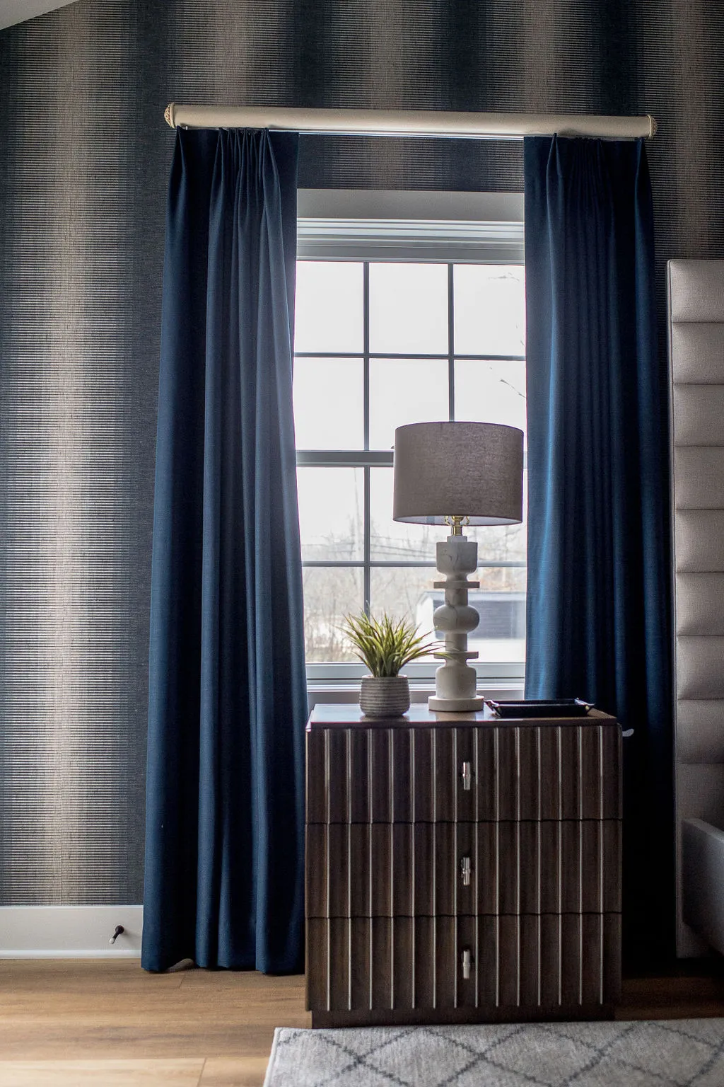 Table lamp sitting on top of a dark wooden nightstand in front of a window with blue panel drapes