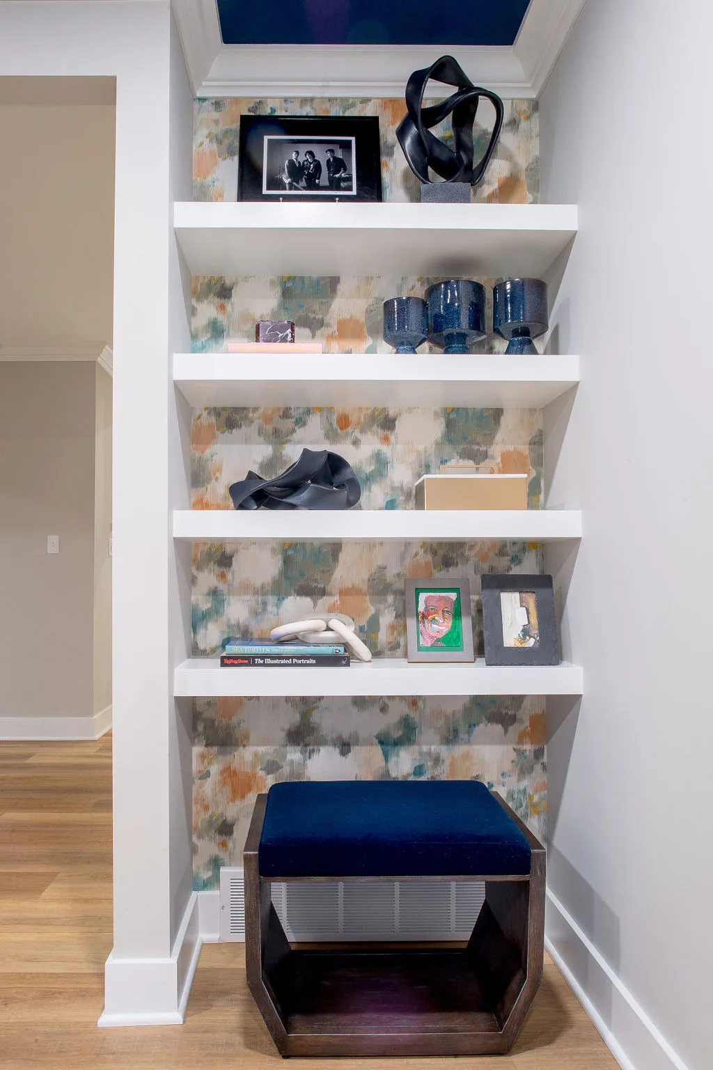 White open shelves in a niche with colorful wallpaper behind, accessories scattered on the shelves, and an ottoman sitting at the bottom