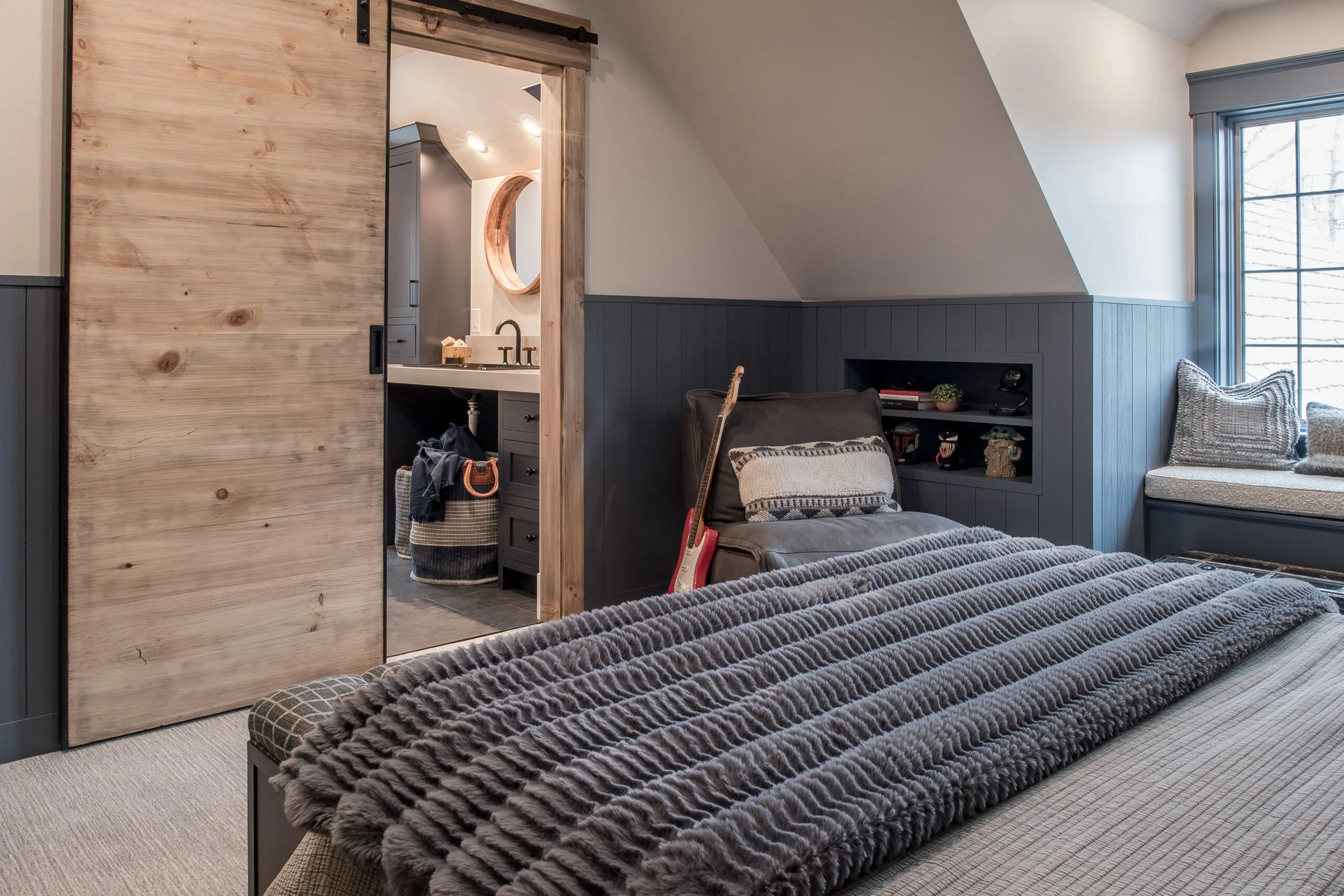 Image in a bedroom looking at the end of a bed with a navy throw blanket and a side chair with a guitar leaning against it in the background
