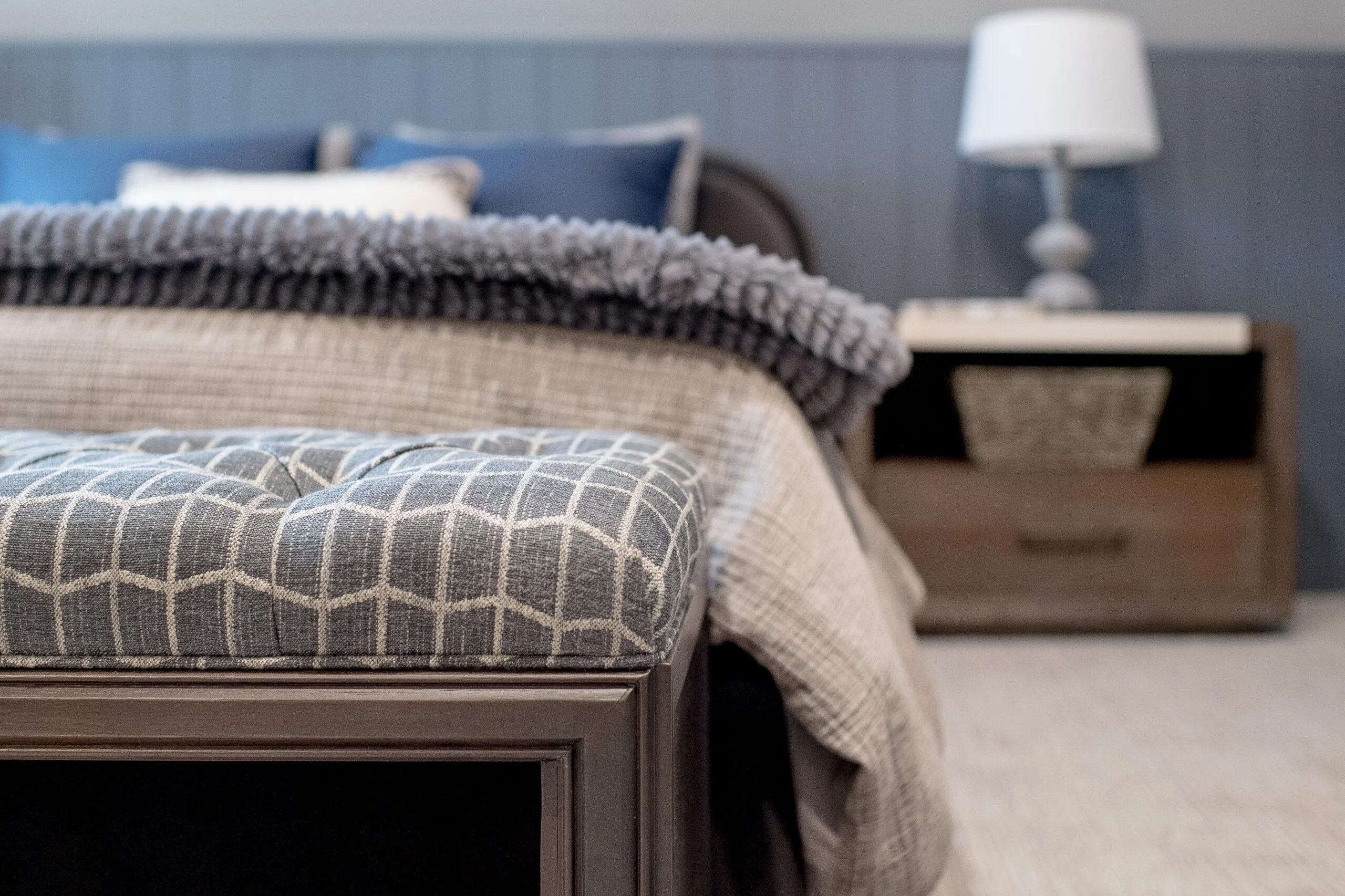Close up image of an upholstered bench at the end of a bed with a side table and lamp in the background