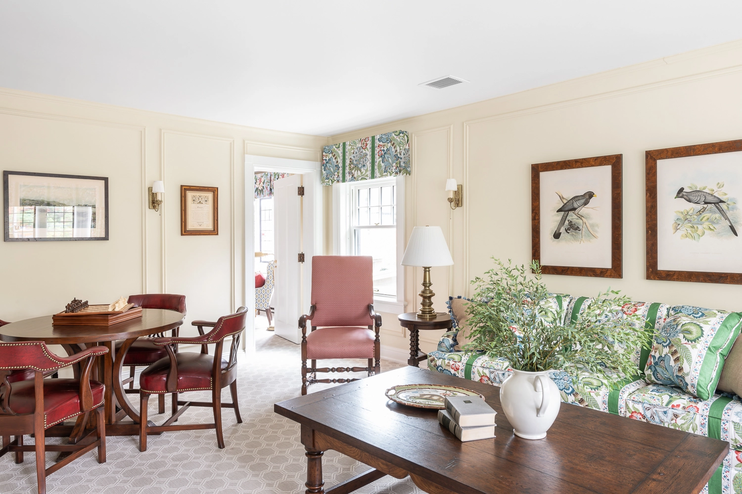 Image of wooden coffee table with books and florals with a sitting table and upholstered red chair in the background