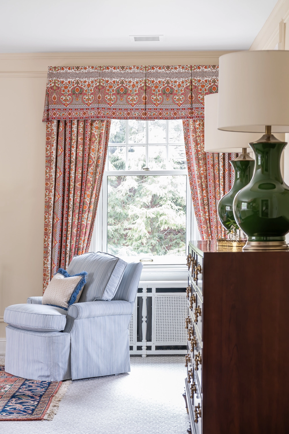 Image of the side of a bedroom dresser with two green lamps, a window and upholstered chair behind
