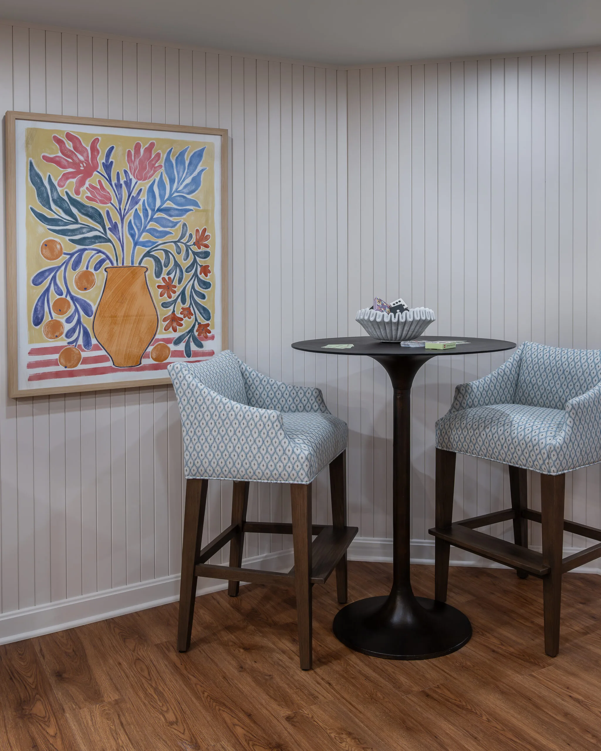 lower level bar high top table with blue upholstered stools and fun floral, colorful artwork