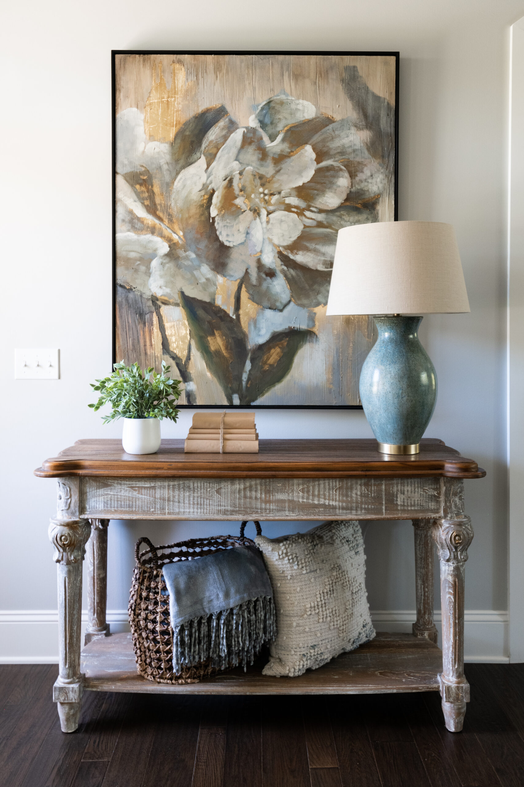An entryway distressed console table with wood top decorated with a blue based lamp with a cream shade. A photo of a flower hangs above in blue hues with gold accents.