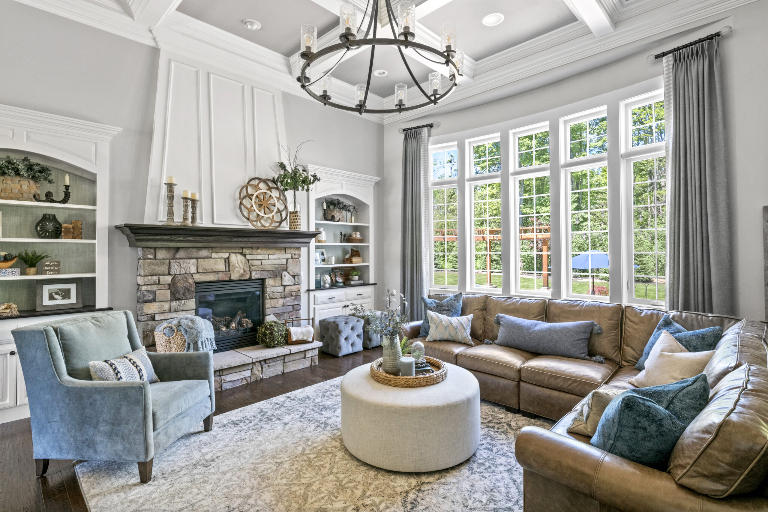 A view of the living room as you walk in from the kitchen with a large window, a stone fireplace, brown couch, circle ottoman, and blue accent pieces